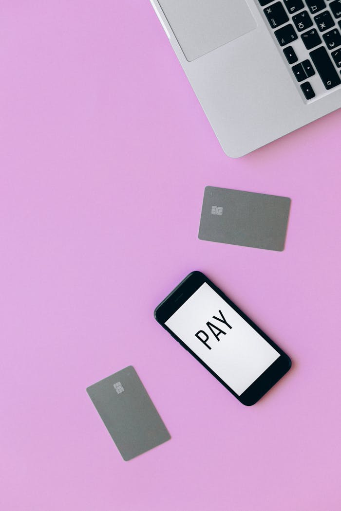 Flat lay of a laptop, smartphone displaying 'PAY', and credit cards on a pink surface representing online transaction.